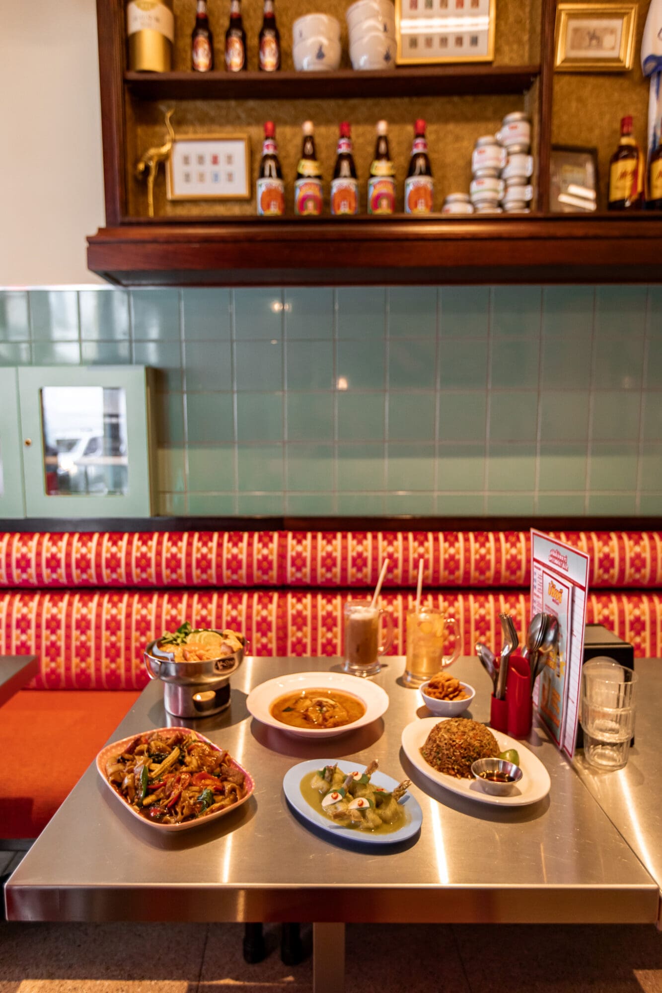 A selection of dishes on a metal table against a red booth and green tiled wall at Speedboat Bar Thai Restaurant.