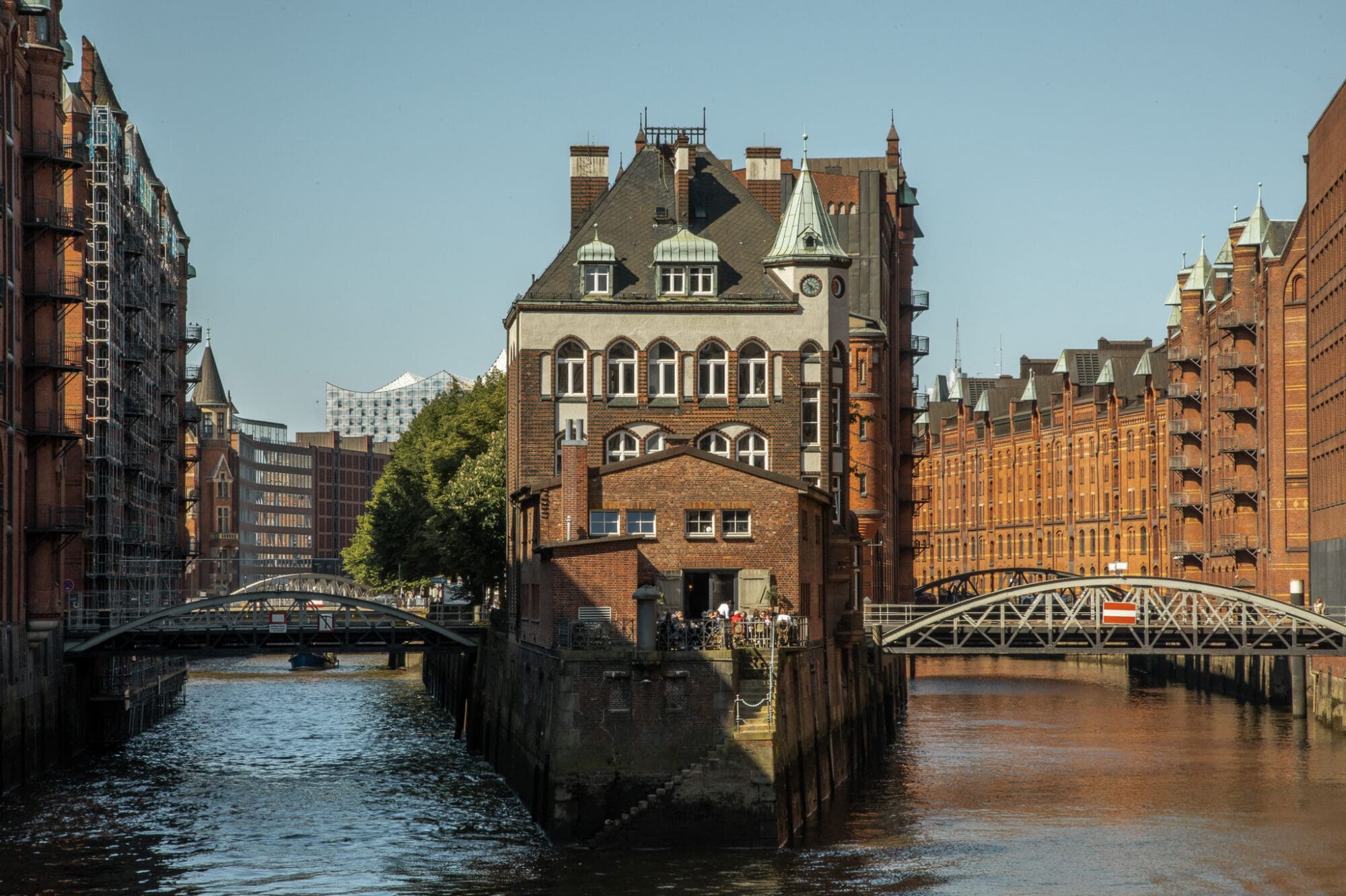 A photograph of a waterway in Hamburg with a brown building in the middle, flanked by two bridges