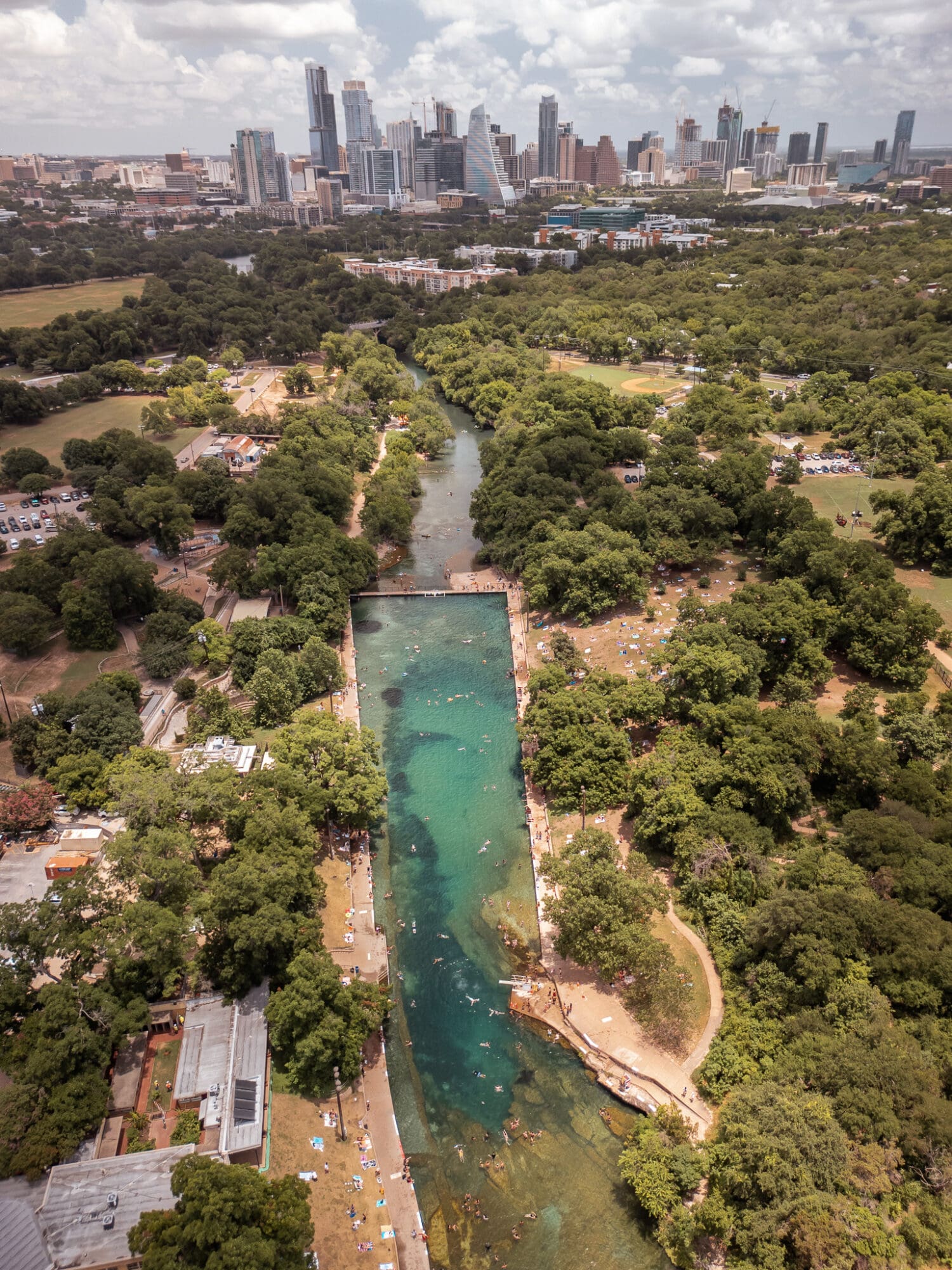 The image is an aerial view of Barton Springs Pool in Austin, Texas. The natural swimming pool is surrounded by lush green trees and grassy areas where people are relaxing and sunbathing. Swimmers can be seen enjoying the clear, turquoise water. The pool is situated within Zilker Park, with paths and small buildings around it. In the background, the downtown Austin skyline is visible, featuring modern high-rise buildings under a partly cloudy sky.