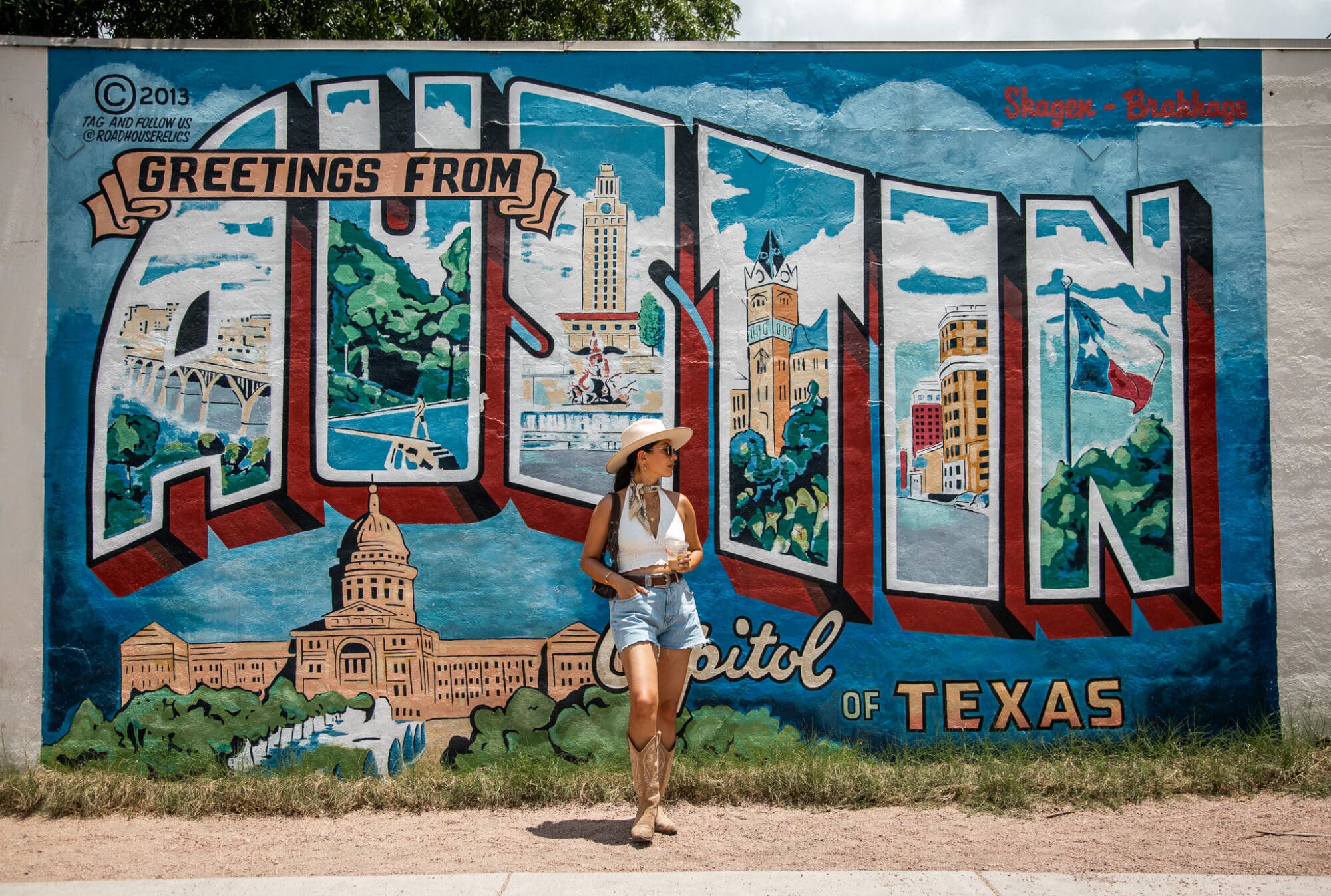 The image shows Anoushka standing in front of a colorful mural that reads "Greetings from Austin, Capitol of Texas." The mural features various iconic images associated with Austin, including the Texas State Capitol building, the University of Texas Tower, and the Texas flag. Anoushka is dressed in a white sleeveless top, denim shorts, a wide-brimmed hat, and cowboy boots.