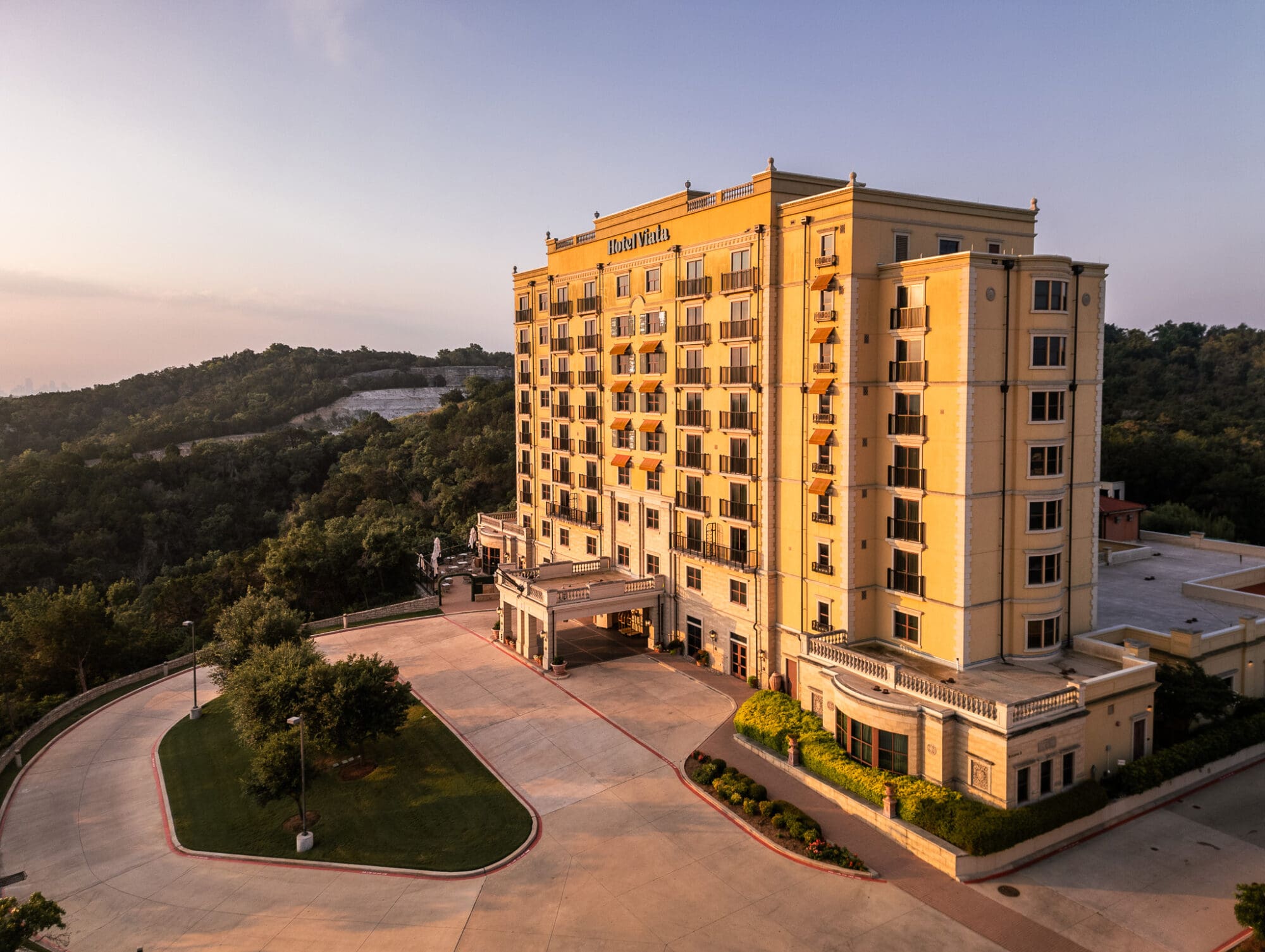 The image is an aerial view of Hotel Viata in Austin, Texas, taken at sunrise. The hotel is a large, elegant building with multiple stories, featuring balconies and large windows. It has a light yellow exterior with decorative elements and a welcoming entrance area. The surrounding landscape consists of rolling hills covered in dense greenery.