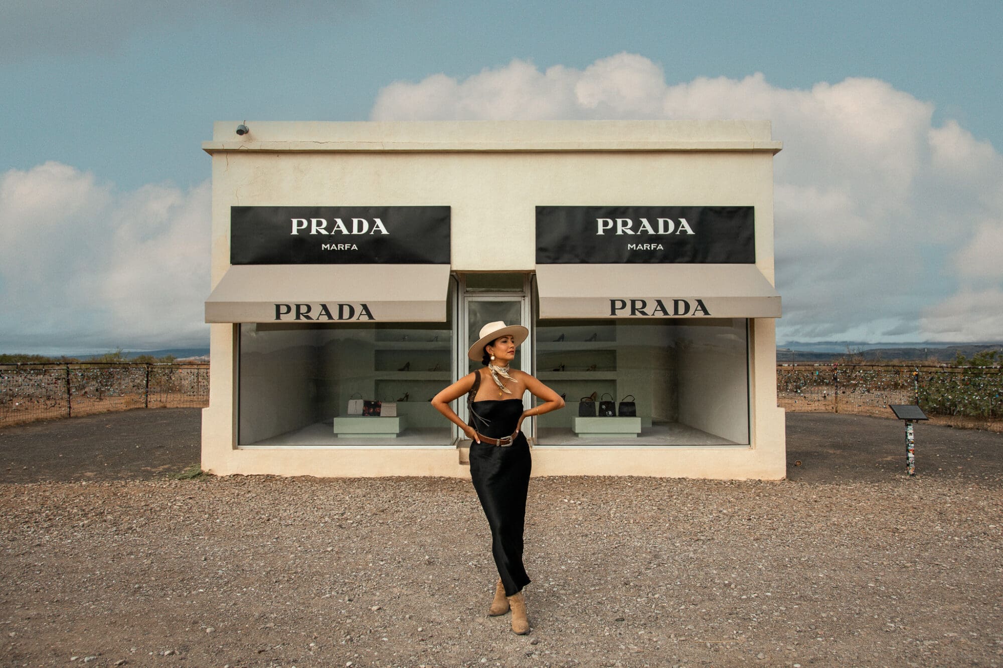 Anoushka stands wearing a black dress, cream hat, and cowboy boots with her hands on her hips. Behind her is Prada Marfa, an art installation replicating a Prada store, placed in the Texan desert.