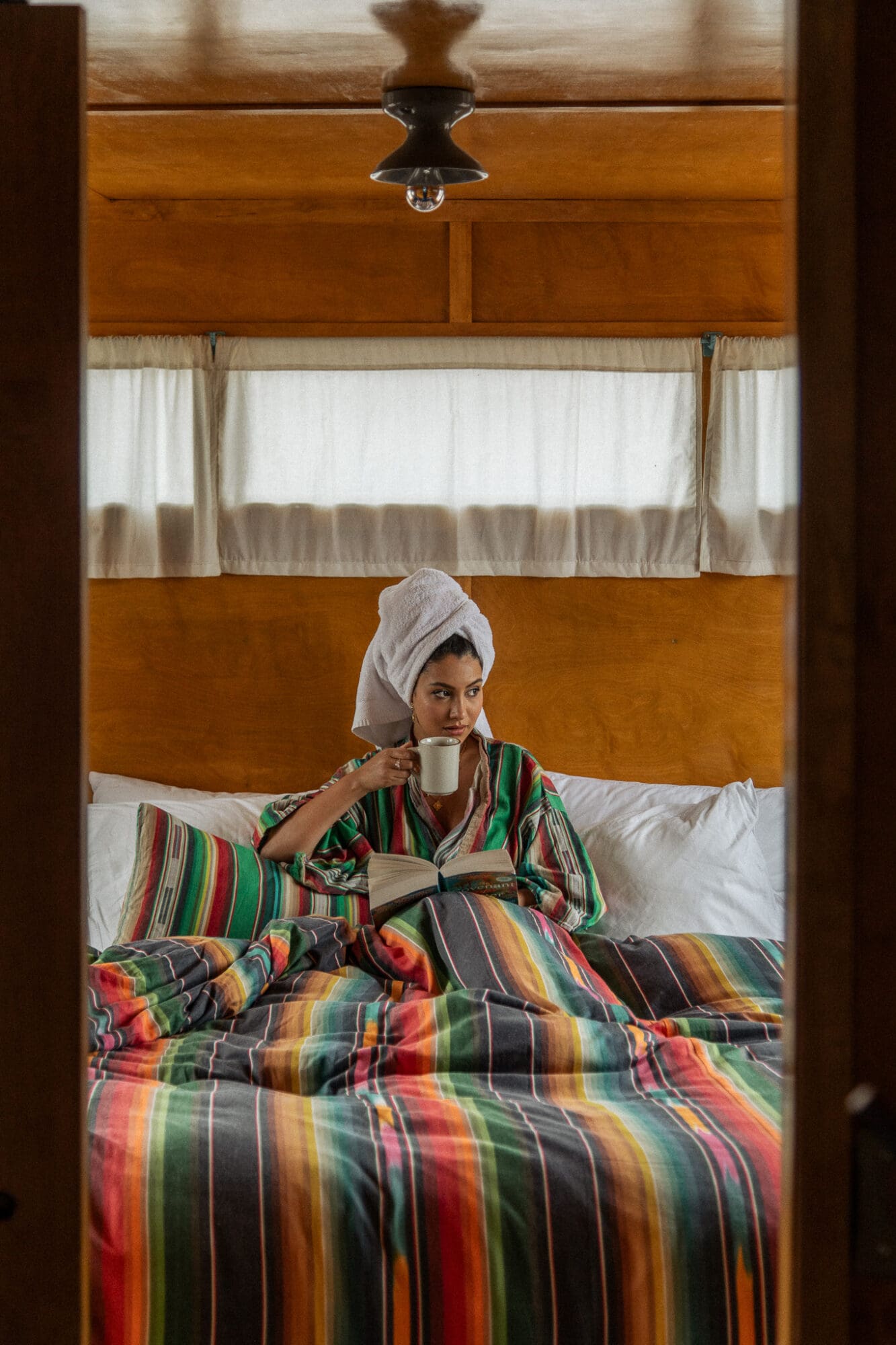 Anoushka sits in bed at her trailer in El Cosmico, Marfa, drinking morning coffee with her hair wrapped in a towel, wearing a multi-coloured striped robe that matches the bed sheets.