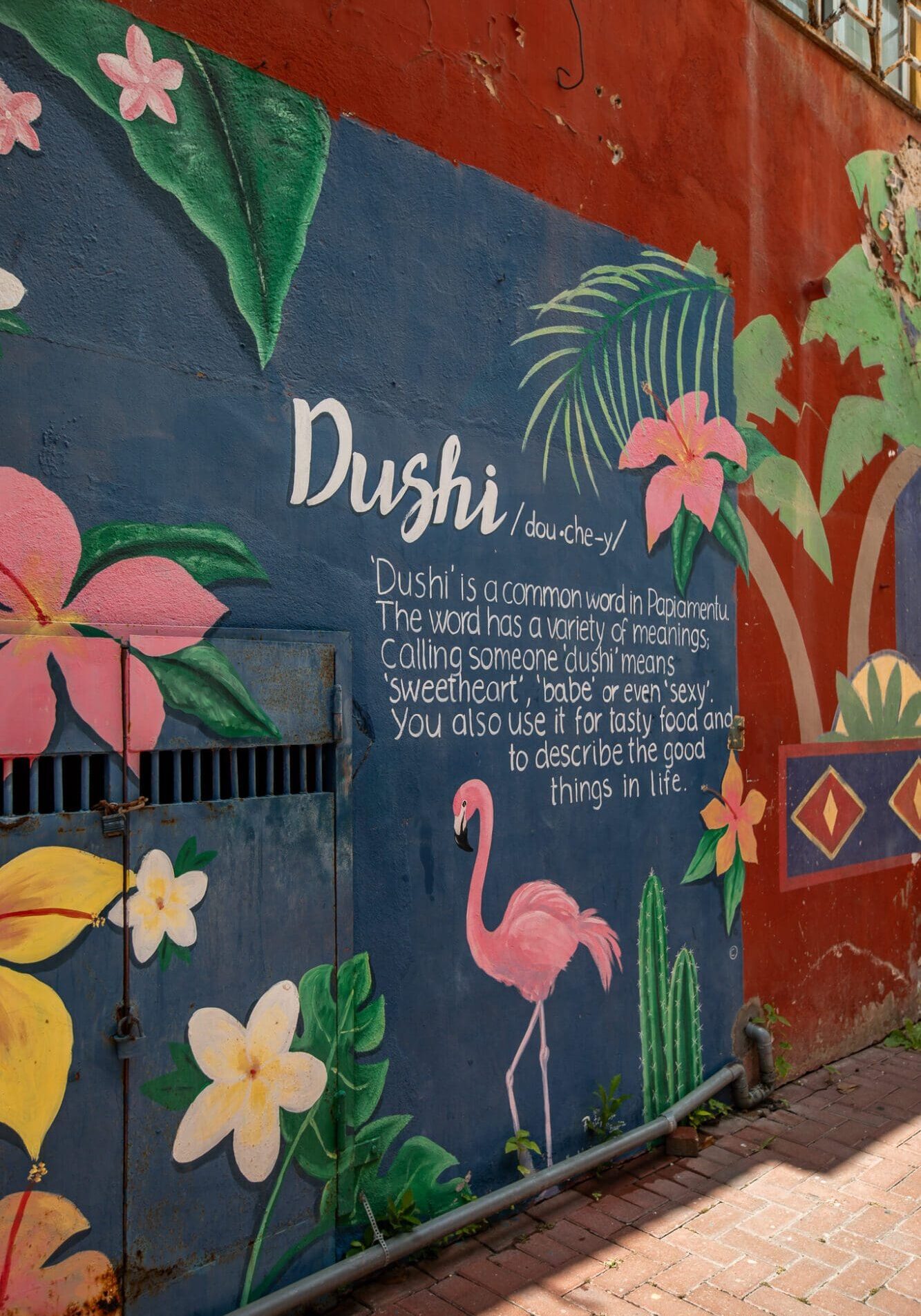 A photograph of a colourful street art mural featuring flamingos, and text explaining the meaning of the word "Dushi", on a wall in Curacao