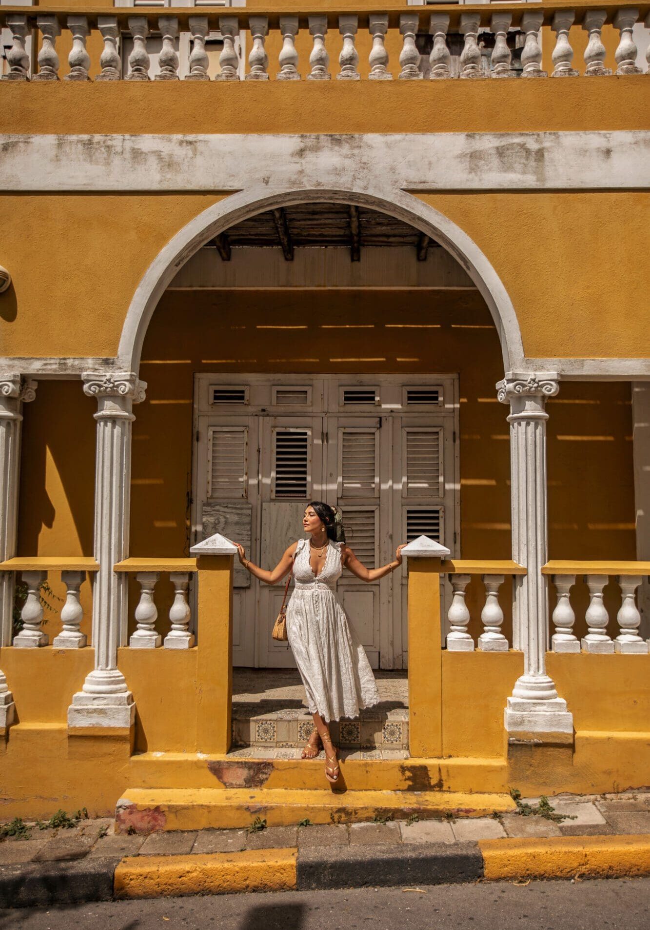 A girl in a white dress looking out from a yellow building with columns in Curacao, The Caribbean