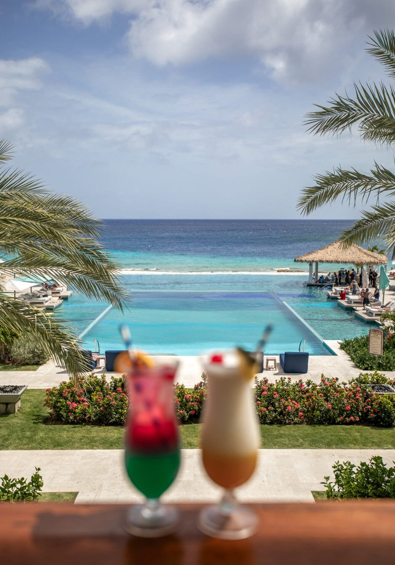 A photo of two colourful cocktails with a backdrop of a double infinity pool and the ocean at Sandals Royal Curacao