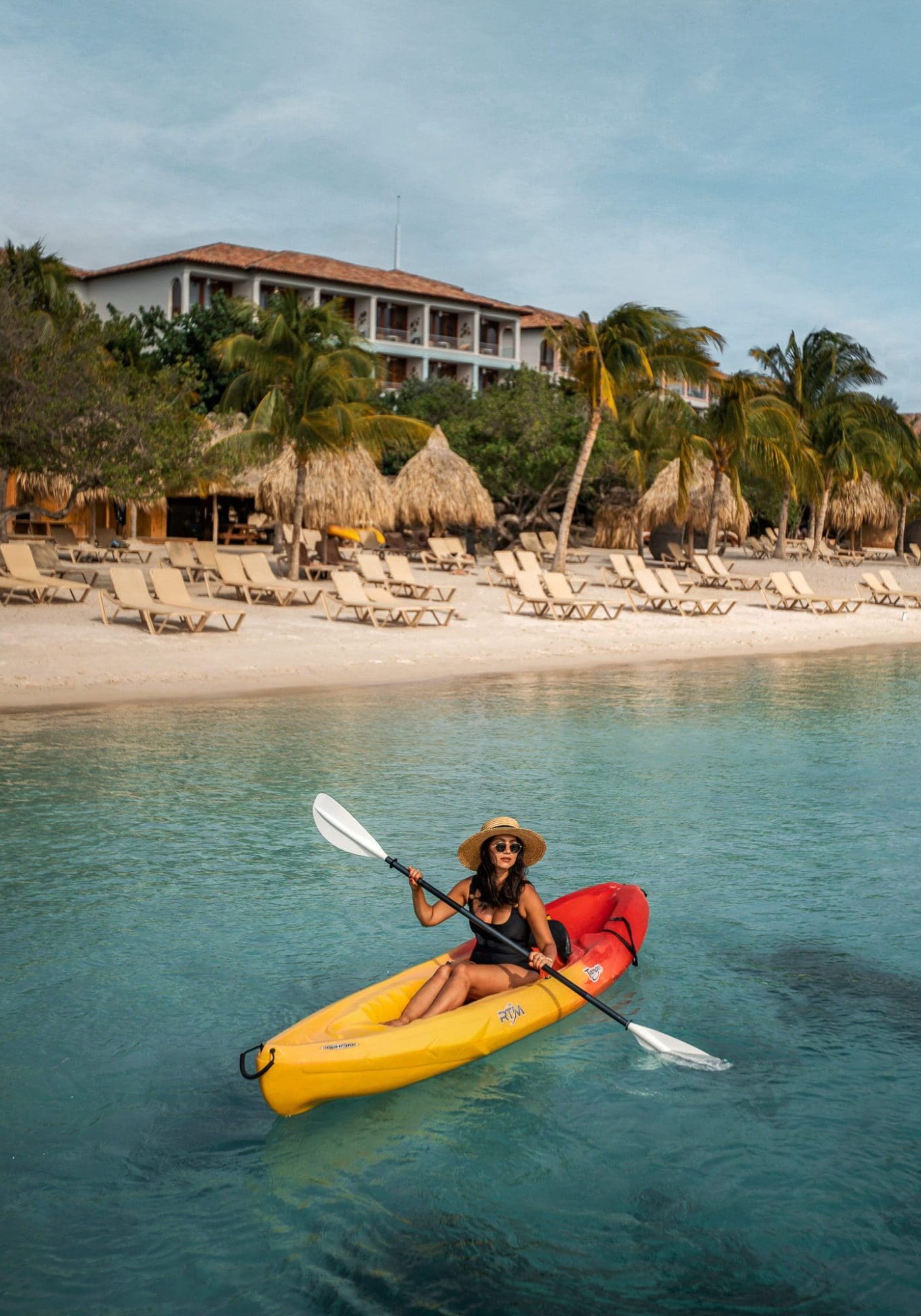 A woman wearing a black swimsuit and sun hat kayaks on a yellow and red kayak through turquoise blue waters in front of the beach at Sandals Royal Curacao