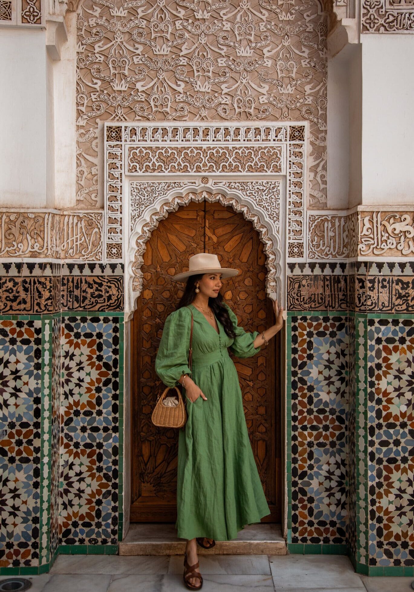 Anoushka stands wearing a green midi dress and fedora hat against an intricate brown door surrounded by green and blue tiles in the Medersa Ben Youssef, Marrakech.