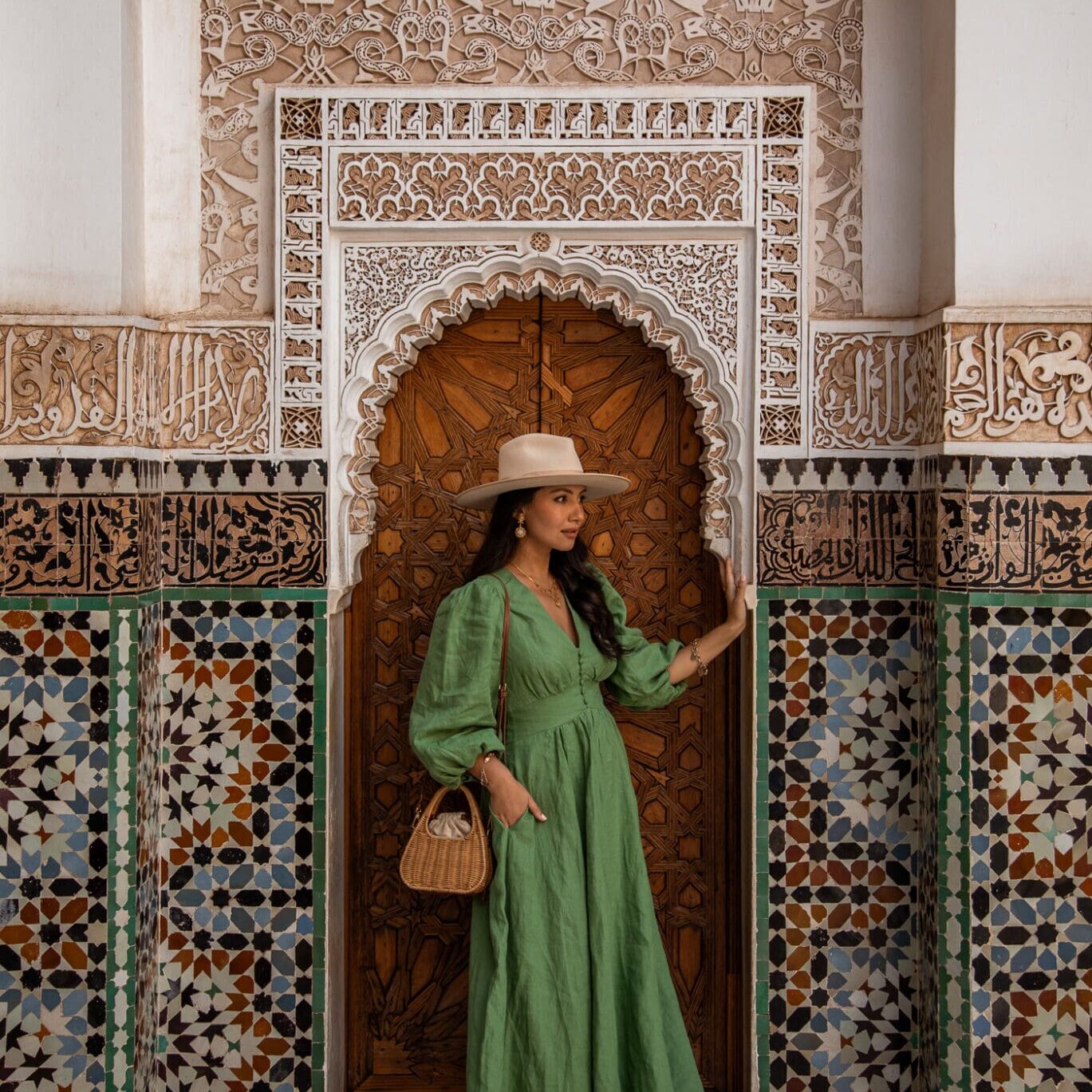 Anoushka stands wearing a green midi dress and fedora hat against an intricate brown door surrounded by green and blue tiles in the Medersa Ben Youssef, Marrakech.