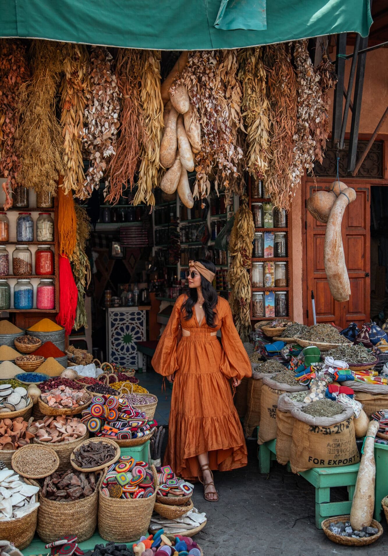 Anoushka wears an orange maxi dress and brown headcarf in the souks of Marrakech, surrounded by colourful pots of spices